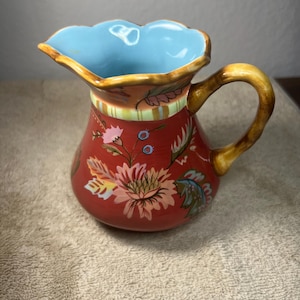 May include: A red ceramic pitcher with a floral design in shades of pink, yellow, and blue. The pitcher has a light blue interior and a brown handle.
