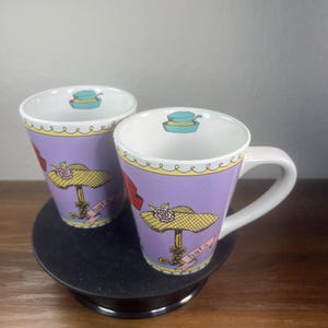 May include: Two ceramic mugs with a lavender exterior featuring a yellow hat illustration with a pink rose and a pink ribbon labeled hats. The interior rim displays a small blue hat graphic. The mugs have a white handle and a scalloped yellow border.