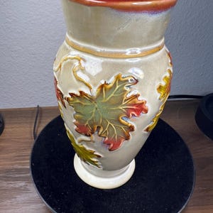 May include: A ceramic vase with a wide opening and a flared rim. The vase is decorated with embossed autumn leaves in shades of green, red, and yellow. The rim has a reddish-brown glaze, and the body is a muted green.