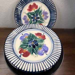 May include: Two round ceramic plates featuring a cream-colored center with a hand-painted floral motif in red, blue, and purple with green leaves. The wide rims are decorated with a dark blue and white striped pattern around the entire edge.