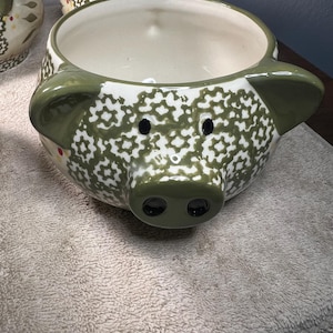 May include: Ceramic pig-shaped bowl with a white interior and green exterior. The bowl features a pig face design with black eyes and green ears. The exterior is decorated with a white and green pattern. A set of bowls is visible.