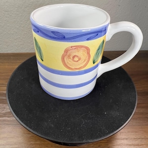 May include: A ceramic mug with a white interior and handle. The exterior features a hand-painted design with a blue wavy line at the top, a yellow band with a central orange flower and green leaves, and two horizontal blue stripes at the base.