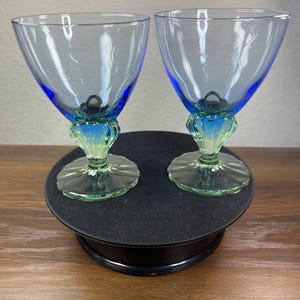 May include: Two matching stemmed glasses featuring a wide, light blue bowl that transitions into a vibrant blue mid-section, ending in a pale green, fluted base. The glass bowls display a subtle, etched floral pattern on the exterior surface.