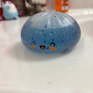 Puede incluir: Una bola antiestrés azul en forma de dumpling con una cara sonriente y detalles de purpurina. La bola antiestrés está sobre una superficie blanca. El dumpling es de color azul claro con purpurina azul oscuro y verde.