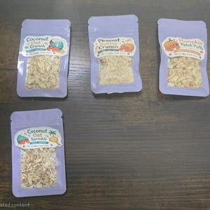 May include: Four light purple resealable pouches containing pet treats. Labels read Coconut Oat Crunch, Peanut Butter Crunch, Pumpkin Patch Puffs, and Coconut Oat Sprinkle. Each pouch features a clear window displaying the dry, textured contents.