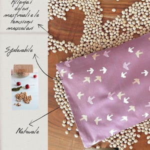 May include: A lavender-coloured, rectangular heating pad with a pattern of white birds. The pad is surrounded by cherry pits and scattered on a wooden surface. Text in Italian reads "Cuscino Termico in noccioli di ciliegia."