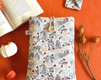 May include: A fabric book sleeve with a botanical pattern in white, green, burgundy, and tan. The sleeve has a wooden button closure and a matching small square item. The background is orange, with a book and decorative elements.