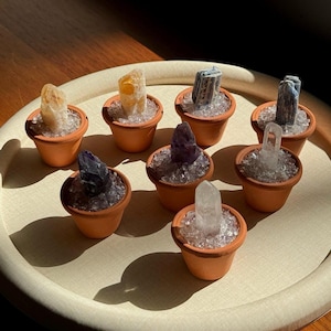 May include: A collection of small terracotta pots, each filled with clear crystals and a variety of colorful gemstones. The pots are arranged on a round, light-colored tray, illuminated by sunlight.
