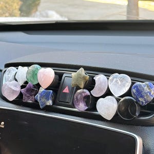 May include: A collection of various crystal heart, star, and moon shapes in different colors, including pink, green, purple, blue, and white, are displayed on a car's dashboard vent.