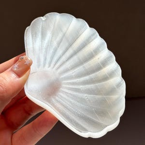 May include: A translucent white shell-shaped dish with a smooth, pearlescent finish and radiating ridges. The object features a scalloped edge and a curved, concave form, reflecting light across its surface with a subtle, shimmering, crystalline texture.