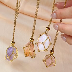 May include: Four gemstone necklaces with gold-colored chains. The gemstones are amethyst, citrine, a white stone, and rose quartz. Each stone is held in a gold-colored wire cage. The background is a light peach color.