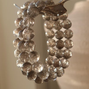 Könnte beinhalten: Ein transparentes Armband mit mehreren Strängen aus runden, glitzergefüllten Perlen. Das Armband wird von einem silbernen Löffel gehalten. Die Perlen haben einen silbernen Glitzereffekt, der ein schimmerndes Aussehen erzeugt. Es ist ein Schmuckstück.