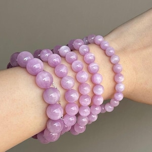 May include: A stack of six lavender-colored gemstone bracelets with round beads. The bracelets are layered on a person's wrist.
