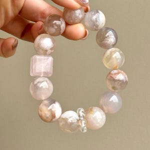 May include: A beaded bracelet featuring smooth, polished, translucent spheres in shades of pale pink, cream, and soft gray. The bracelet includes one square-shaped bead and a small, clear, faceted spacer bead among the round, marbled stones.