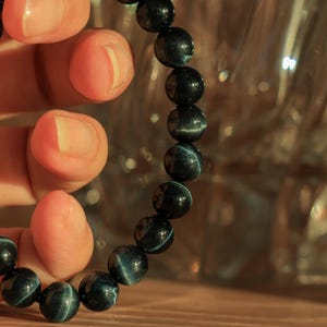 May include: A dark blue beaded bracelet with a tiger's eye pattern. The bracelet is made of round beads and is being held by a hand.