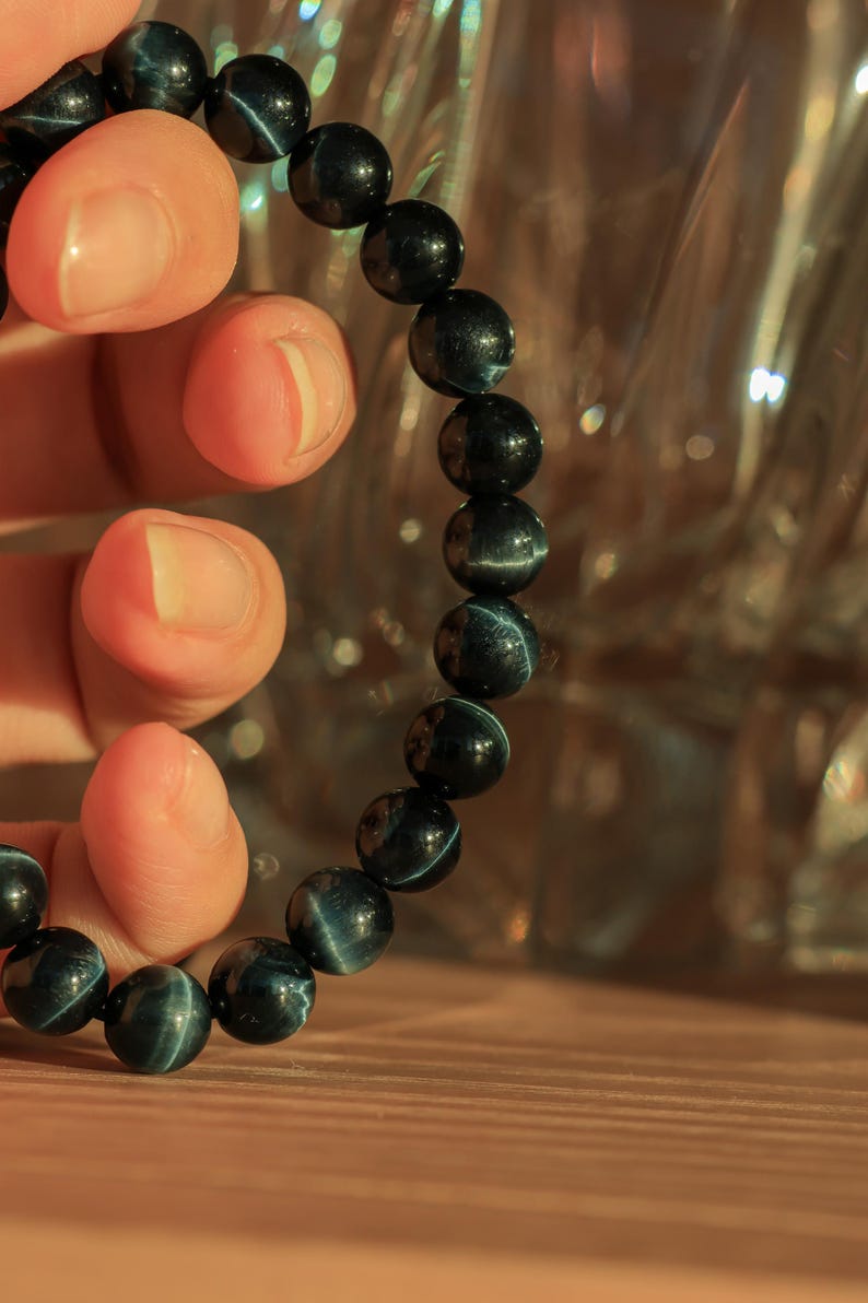 May include: A dark blue beaded bracelet with a tiger's eye pattern. The bracelet is made of round beads and is being held by a hand.