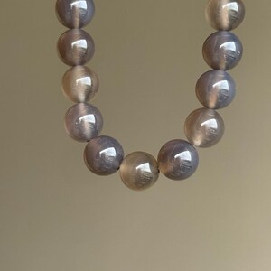 May include: A necklace composed of large, round, polished beads. The beads have a translucent, pearlescent quality with a mix of gray and tan tones. The necklace is displayed against a neutral background.