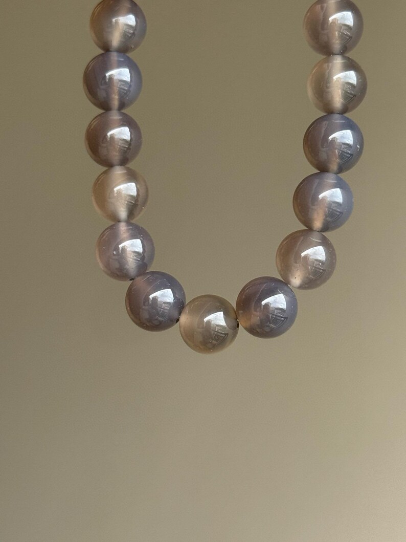 May include: A necklace composed of large, round, polished beads. The beads have a translucent, pearlescent quality with a mix of gray and tan tones. The necklace is displayed against a neutral background.