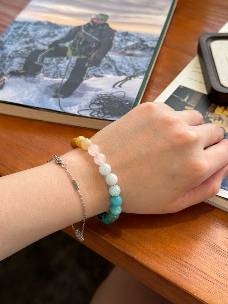 May include: A colorful beaded bracelet with a gradient of yellow, pink, light blue, and turquoise beads. The bracelet is on a person's wrist.