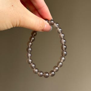 May include: A bracelet made of translucent, smoky gray beads. The beads are round and strung together, forming a circular shape. The bracelet is held by a hand against a neutral background.