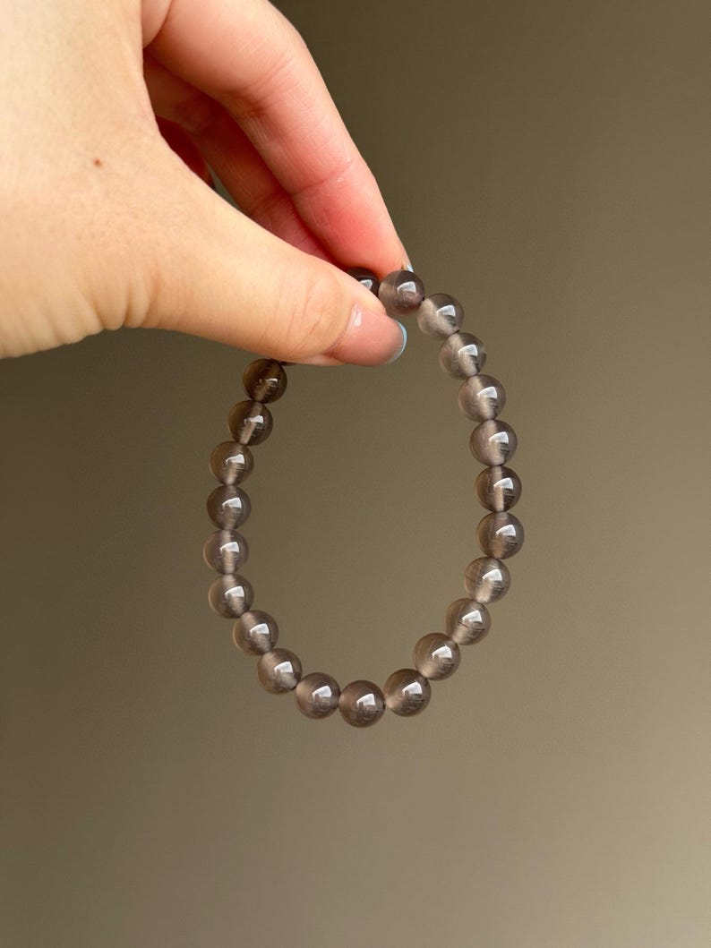 May include: A bracelet made of translucent, smoky gray beads. The beads are round and strung together, forming a circular shape. The bracelet is held by a hand against a neutral background.