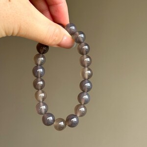 May include: A bracelet made of round, gray-brown beads. The beads have a polished, reflective surface. The bracelet is held up against a neutral background. The beads appear to be made of a natural stone.