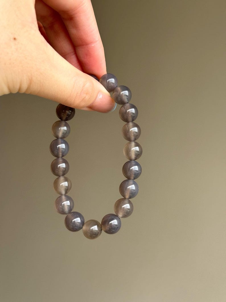 May include: A bracelet made of round, gray-brown beads. The beads have a polished, reflective surface. The bracelet is held up against a neutral background. The beads appear to be made of a natural stone.