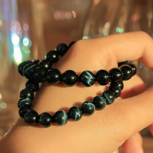 May include: Two black beaded bracelets with blue tiger's eye stones. The bracelets are stacked on a person's wrist.