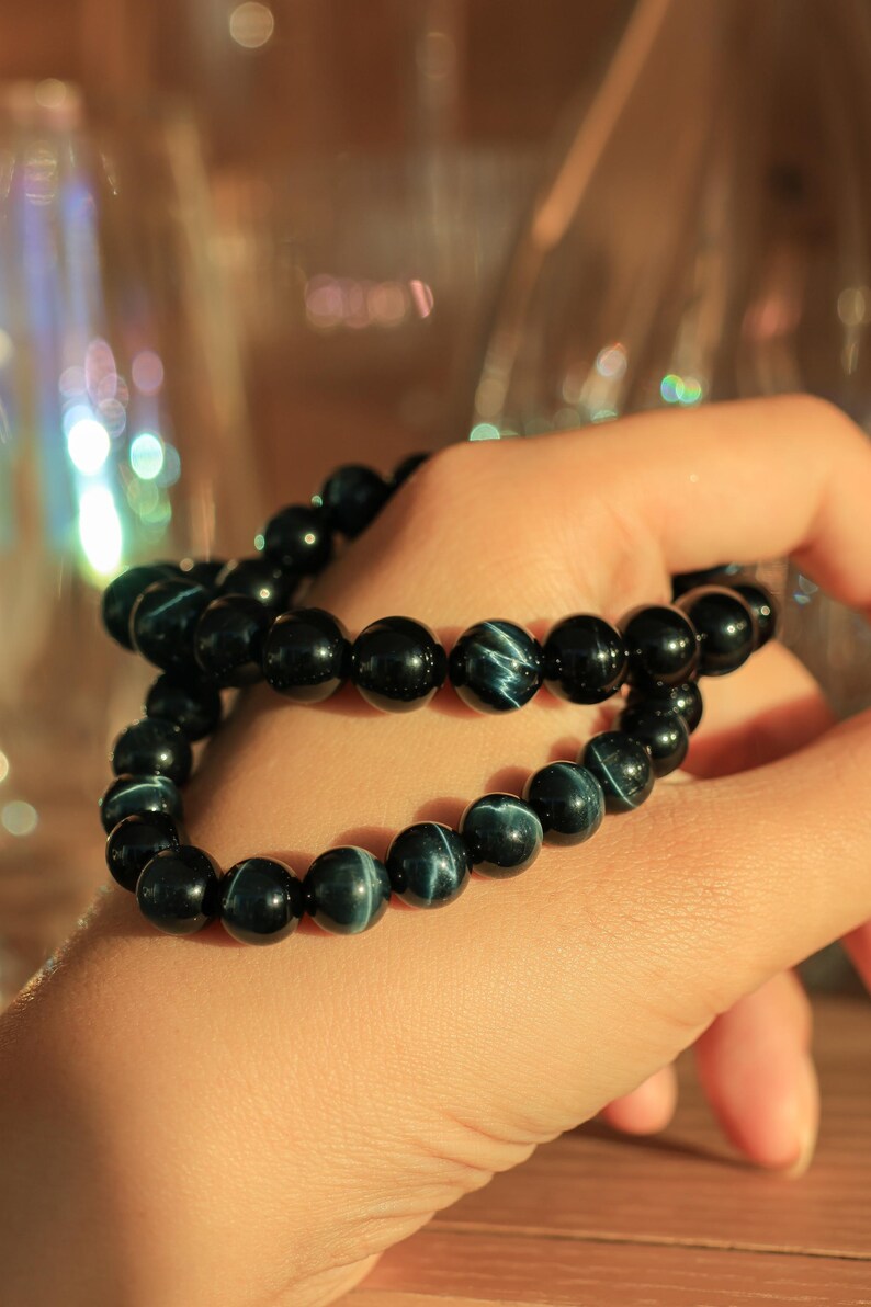 May include: Two black beaded bracelets with blue tiger's eye stones. The bracelets are stacked on a person's wrist.