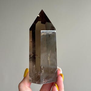 May include: A polished smoky quartz crystal tower with a hexagonal shape and a pointed top. The crystal exhibits a translucent, brownish-grey color with visible internal inclusions and natural fractures throughout the stone structure.
