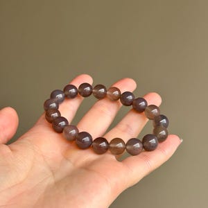 May include: A bracelet made of round, polished, gray-brown beads. The beads are strung together to form a circular shape. The bracelet is held in a hand against a solid, neutral background. The beads appear to be approximately 0.5 inches in diameter.