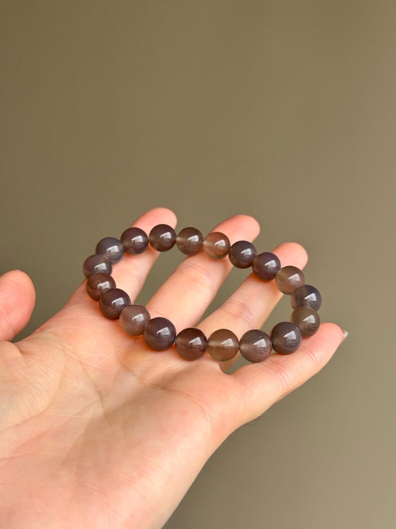 May include: A bracelet made of round, polished, gray-brown beads. The beads are strung together to form a circular shape. The bracelet is held in a hand against a solid, neutral background. The beads appear to be approximately 0.5 inches in diameter.