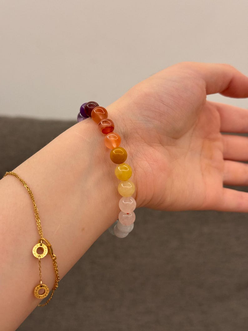 May include: A rainbow colored beaded bracelet with round, smooth stones. The bracelet is worn on a person's wrist.