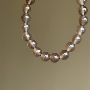 May include: A bracelet featuring a strand of translucent, smoky quartz beads. The round beads are a soft gray color and appear smooth and polished. The bracelet is held up against a neutral background.
