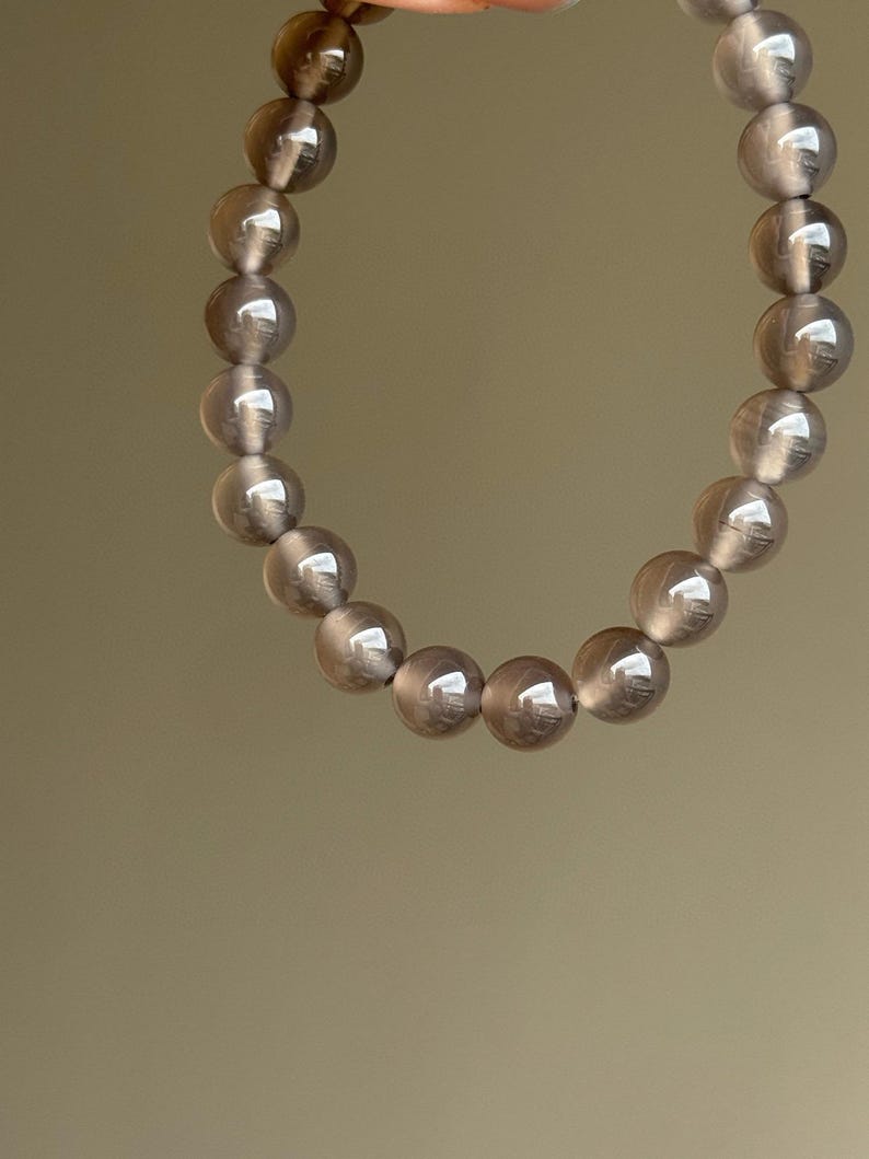 May include: A bracelet featuring a strand of translucent, smoky quartz beads. The round beads are a soft gray color and appear smooth and polished. The bracelet is held up against a neutral background.