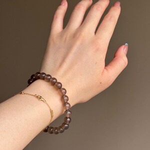 May include: A wrist adorned with two bracelets. One is a beaded bracelet with dark brown, round beads. The other is a delicate gold chain bracelet. The background is a solid, neutral color.