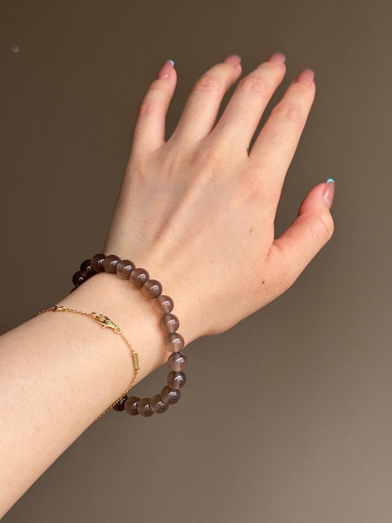 May include: A wrist adorned with two bracelets. One is a beaded bracelet with dark brown, round beads. The other is a delicate gold chain bracelet. The background is a solid, neutral color.