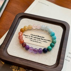 May include: A rainbow colored gemstone bracelet with a wooden box. The bracelet is made of various gemstones, including amethyst, turquoise, and lapis lazuli. The box is made of dark wood and has a light brown fabric lining. The text on the box reads: "Classical Memories Elegance is The only beauty that never fades Hand Craftsmanship and Home Authitics Wooden jewelry box"