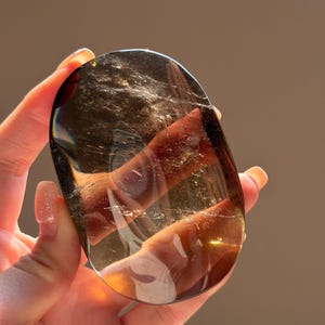 May include: A polished, oval-shaped smoky quartz crystal with a translucent, dark brown hue. The stone features visible internal fractures and natural inclusions that catch the light, creating a reflective, glassy surface with varying depth.