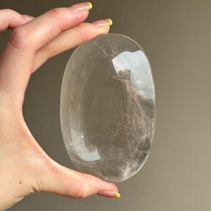 May include: A clear, oval-shaped crystal stone with a polished surface. The stone is held between fingers, showcasing its transparency and subtle internal patterns. The crystal's form is smooth and rounded, reflecting light and creating a sense of depth.