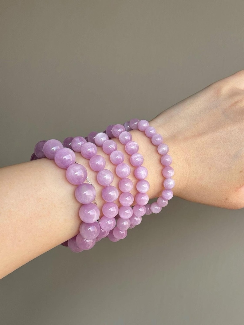 May include: A stack of six lavender-colored gemstone bracelets with round beads. The bracelets are layered on a person's wrist.