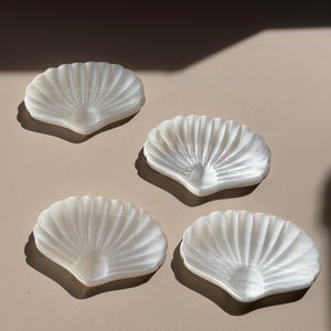 以下が含まれることがあります： グループで配置された4つの白い半透明の貝殻の形をした皿。各皿にはスカラップ状の縁があり、中心点から放射状に広がるリブ状の扇形のテクスチャが施されており、自然のホタテ貝の外観を模しています。