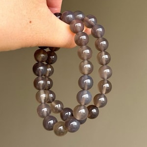 May include: Two bracelets made of gray-brown, translucent, round beads. The beads are strung together, forming a continuous loop. The bracelets are held up against a neutral background.