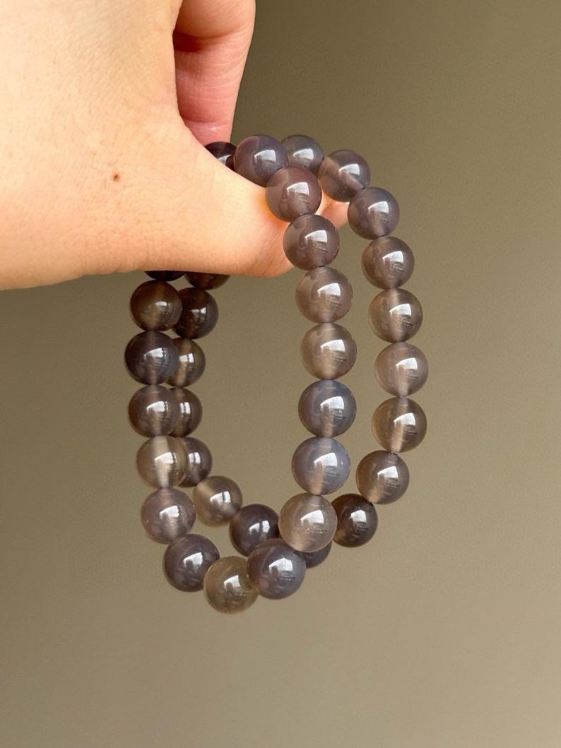 May include: Two bracelets made of gray-brown, translucent, round beads. The beads are strung together, forming a continuous loop. The bracelets are held up against a neutral background.