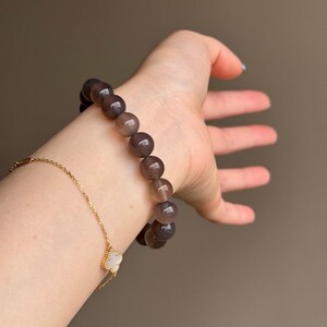 May include: A beaded bracelet with large, round, gray-brown beads. A gold chain bracelet with a clover-shaped charm is also visible. The bracelets are displayed on a person's wrist against a neutral background.
