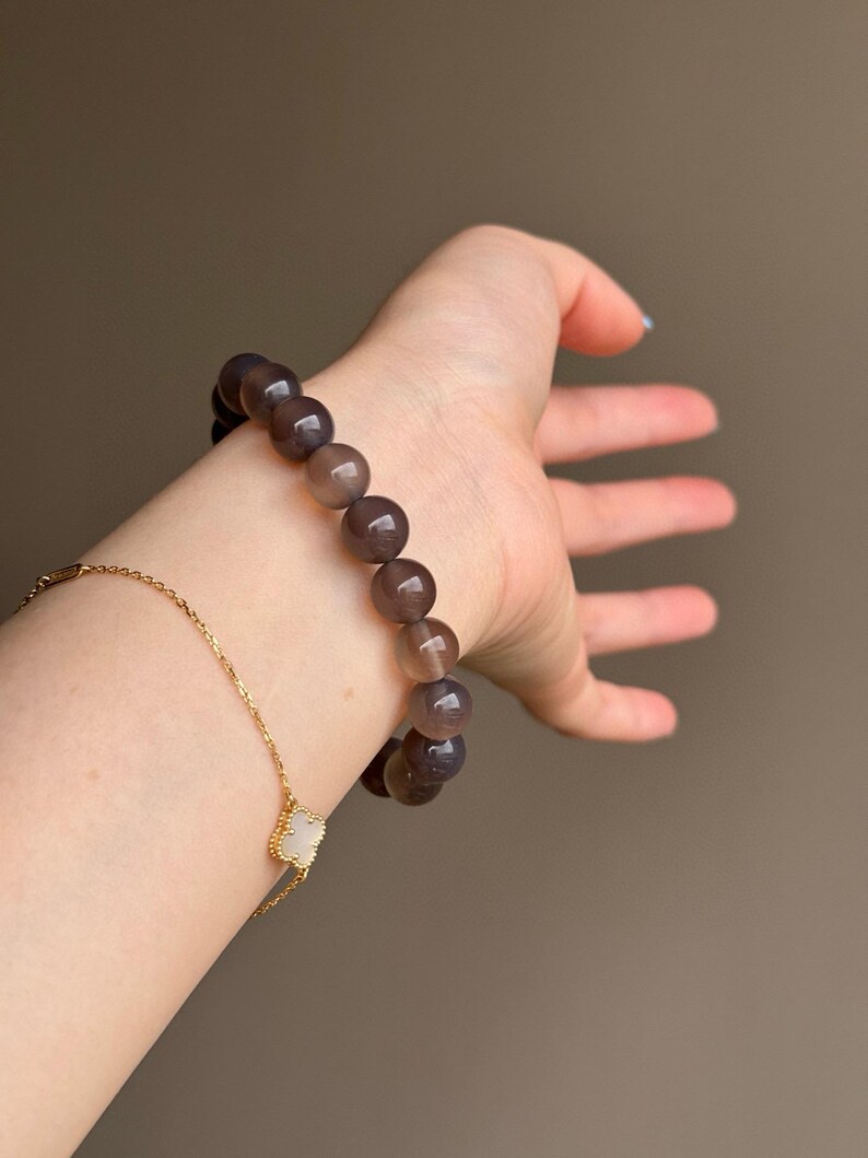May include: A beaded bracelet with large, round, gray-brown beads. A gold chain bracelet with a clover-shaped charm is also visible. The bracelets are displayed on a person's wrist against a neutral background.