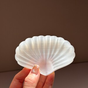 以下が含まれることがあります： 半透明のオフホワイトの貝殻型ディッシュで、扇状の畝のある質感が特徴です。表面は滑らかでわずかに真珠のような光沢があり、光を反射して、湾曲した縁や中央の隆起に繊細なハイライトを作り出しています。