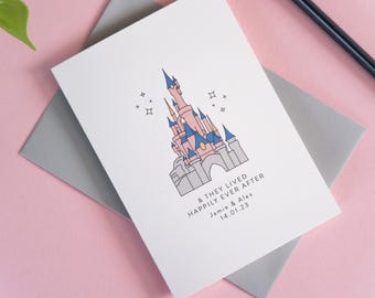May include: A white greeting card featuring a line art illustration of a pink and blue castle with small star accents. Below the castle, the text reads & THEY LIVED HAPPILY EVER AFTER, followed by the names Jamie & Alex and the date 14.01.23.