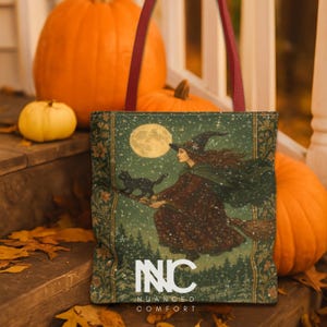 May include: A canvas tote bag featuring a witch on a broomstick with a black cat, a full moon, and a snowy forest scene. The bag has burgundy handles and the text "INC NUANCED COMFORT" at the bottom. Pumpkins and autumn leaves are in the background.