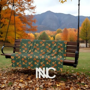 May include: A dark green blanket with a repeating pattern of orange and yellow autumn leaves draped over a wooden swing. The swing is suspended by black chains, set against a backdrop of fall foliage and mountains.